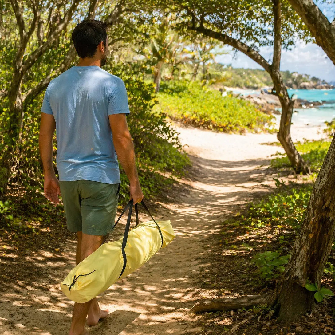 Man walking along a trail with a yellow bag, surrounded by greenery and a beach in the distance.
