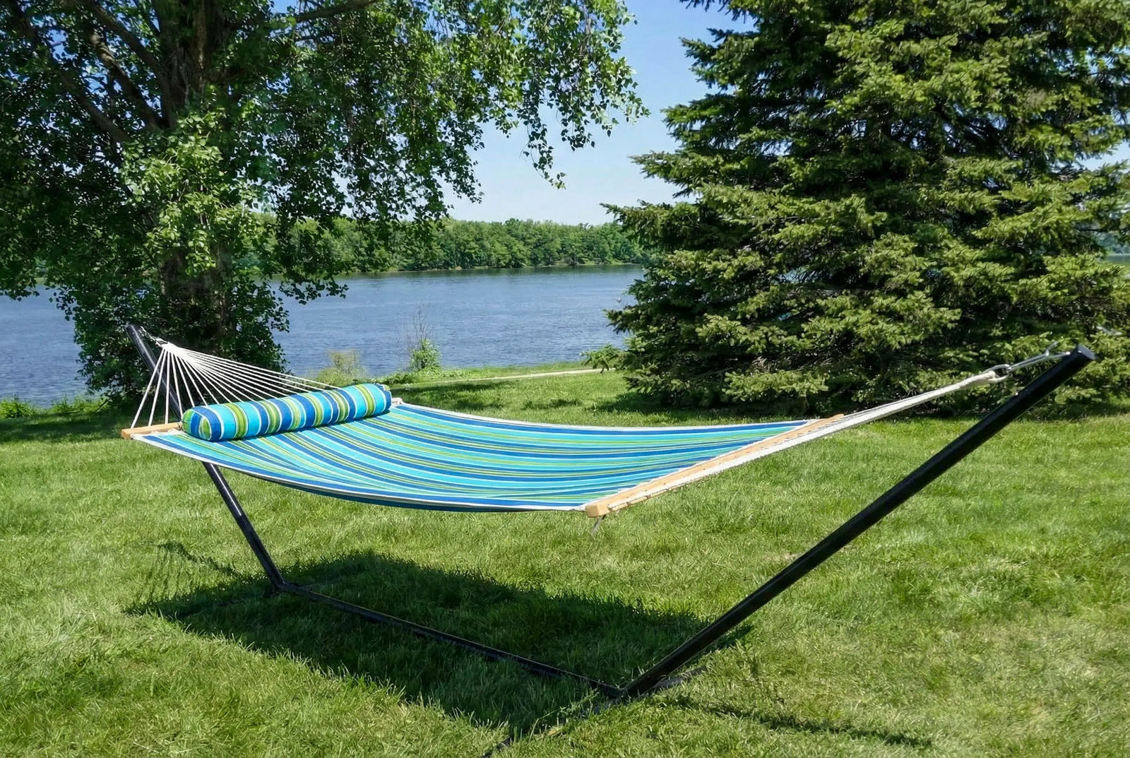 Blue hammock with striped pillow on a stand in a grassy area by a lake. QHD-CAROLINA+15TBSB