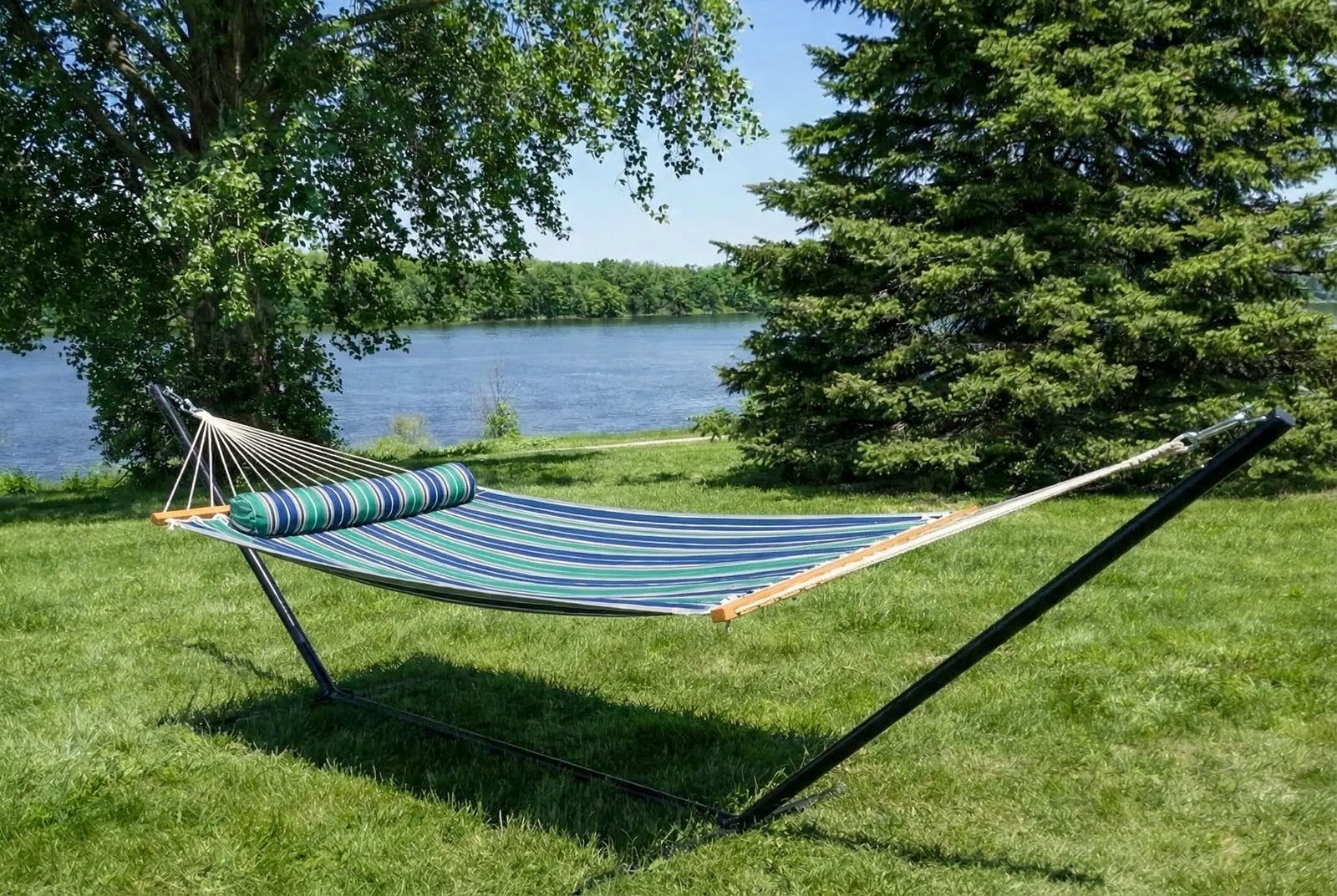 Striped hammock on a stand in a grassy area near a body of water with trees. QHD-GBGW+15TBSB