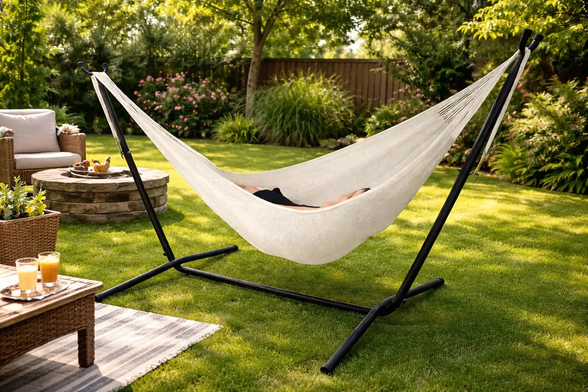 Person relaxing in a hammock on a stand in a garden setting with a fire pit and outdoor furniture.