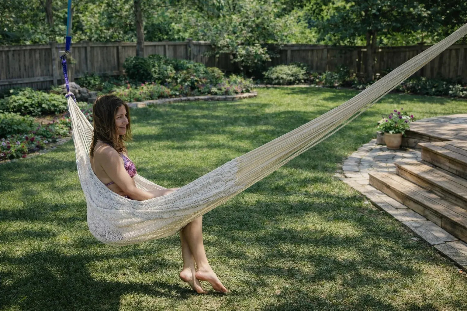 Woman sitting in a hammock in a backyard with green grass and trees. #24-MHXLTC-N