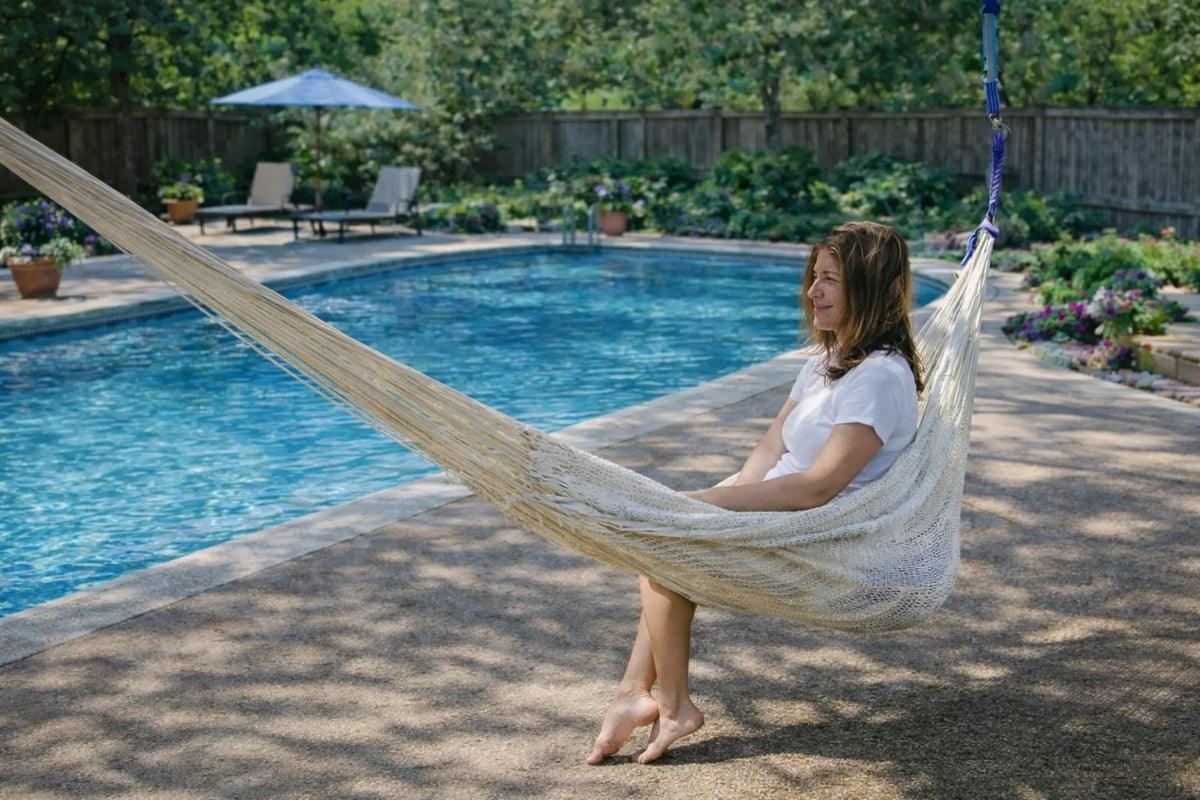 Woman sitting in a hammock by a pool with a garden in the background #24-MHXLTC-N