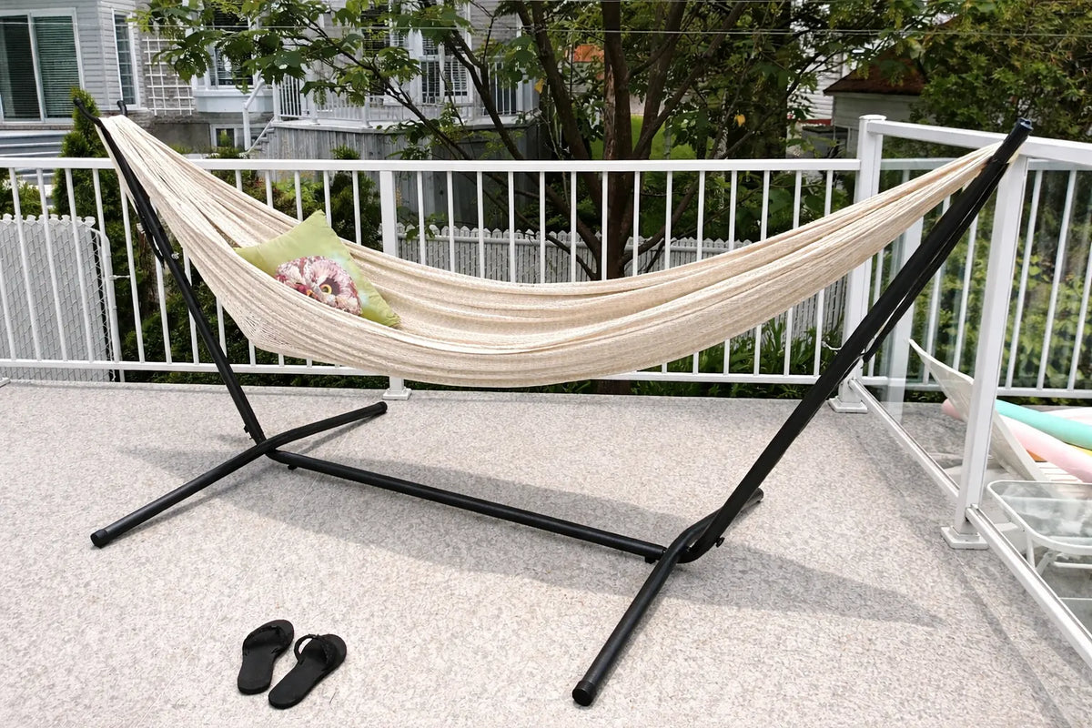 Beige hammock on a black stand on a patio with a white fence and trees in the background.  #24-MHXLTC-N+75121